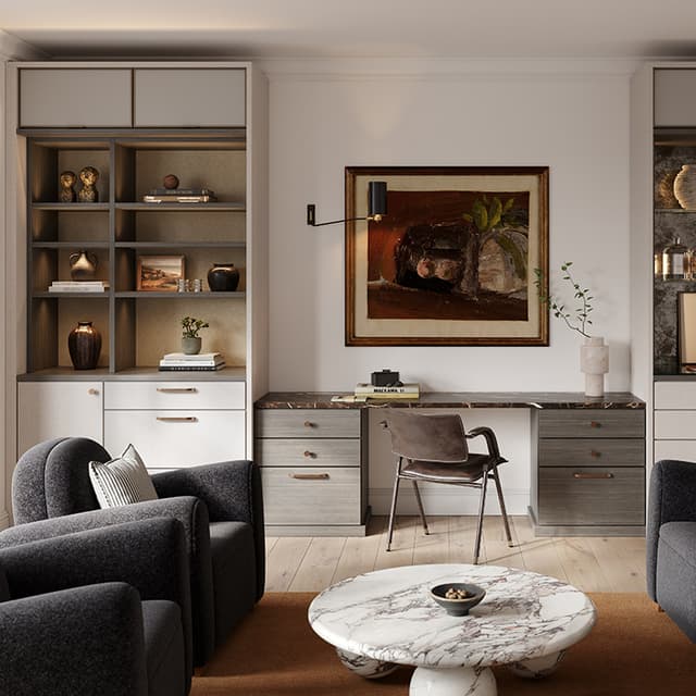 A sleek home office setup with a built-in desk flanked by open shelving, featuring grey drawers, a brown leather chair, and decorative accents like books, vases, and plants. The space includes dark grey armchairs, a marble coffee table, and a large framed artwork above the desk.