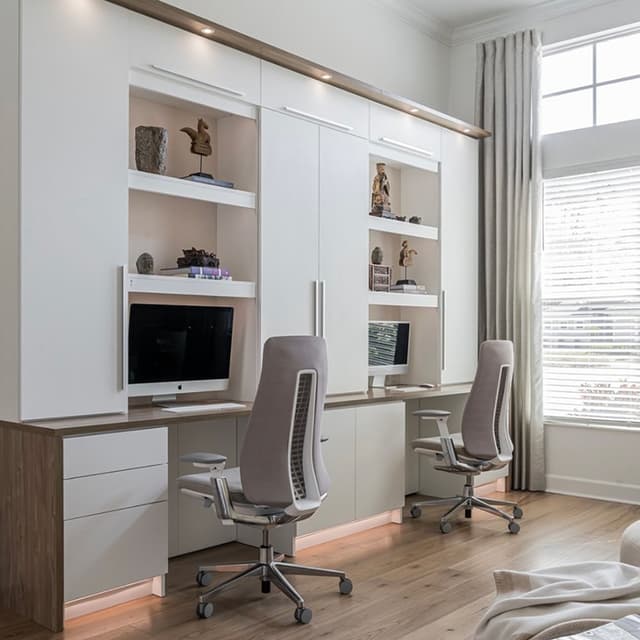 A contemporary home office featuring two workstations with computers, ergonomic chairs, built-in white cabinetry with open shelving, a large window with curtains, and warm wooden flooring.