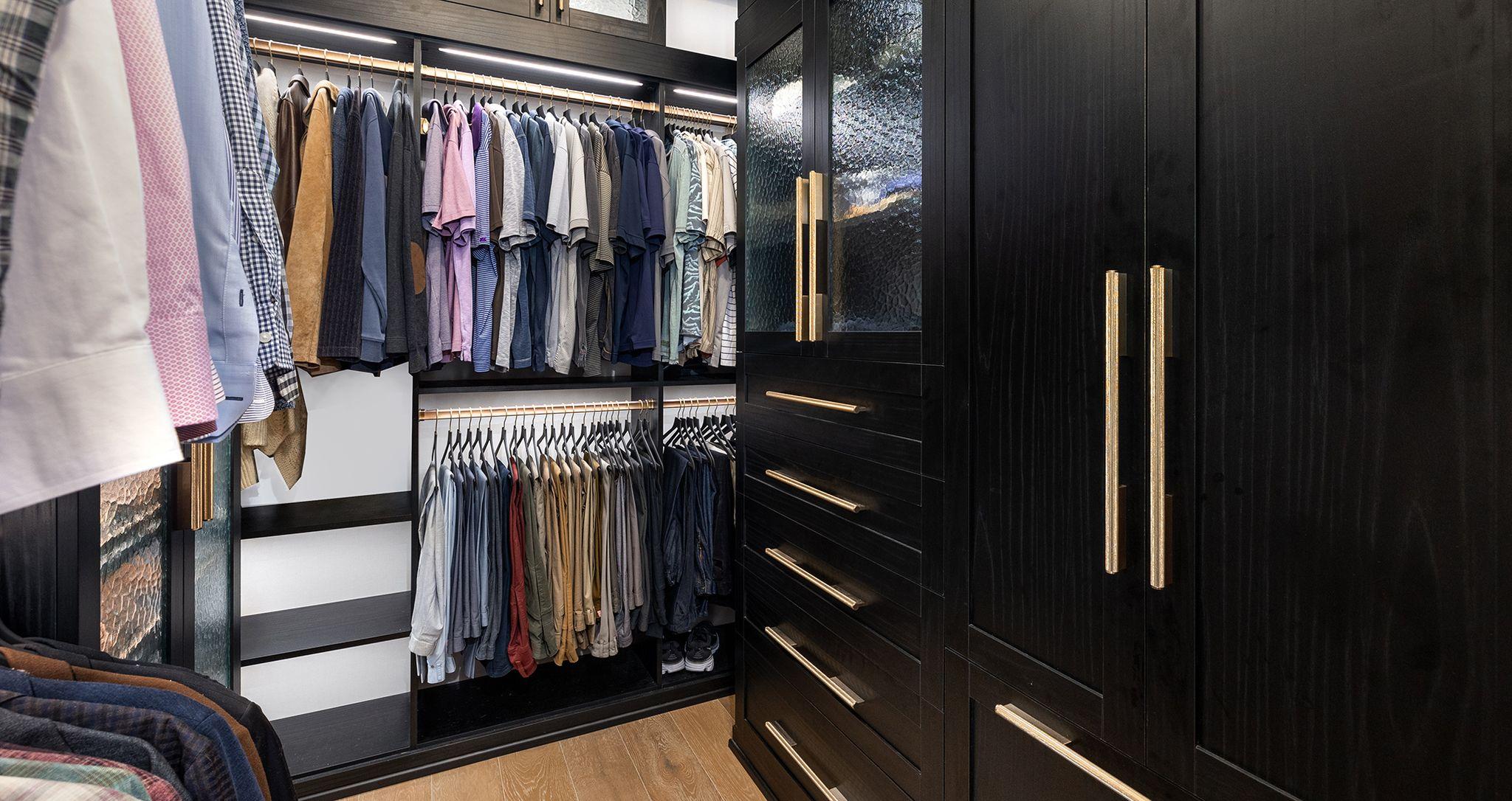 El lado del armario diseñado para él en un acabado de grano de madera oscura con herrajes dorados, estantes personalizados, una cómoda incorporada e iluminación LED de California Closets
