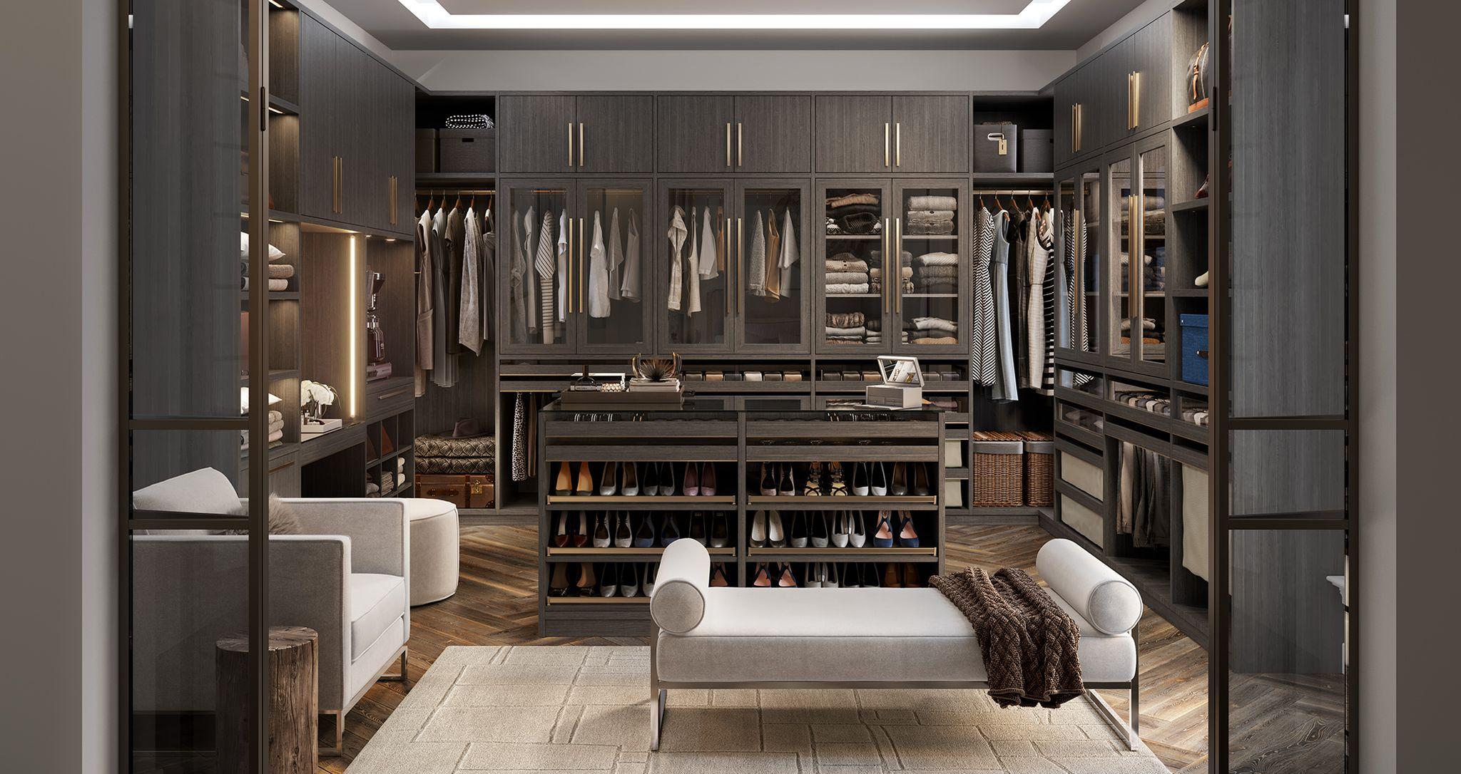 Closet design in u-shape in modern dark wood grain finish with custom shoe storage, glass door cabinets and center seating by California Closets