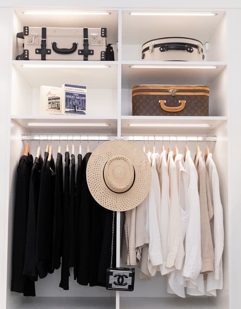 Closet shelving and hanging clothes poles under LED lighting in a white wood grain finish by California Closets