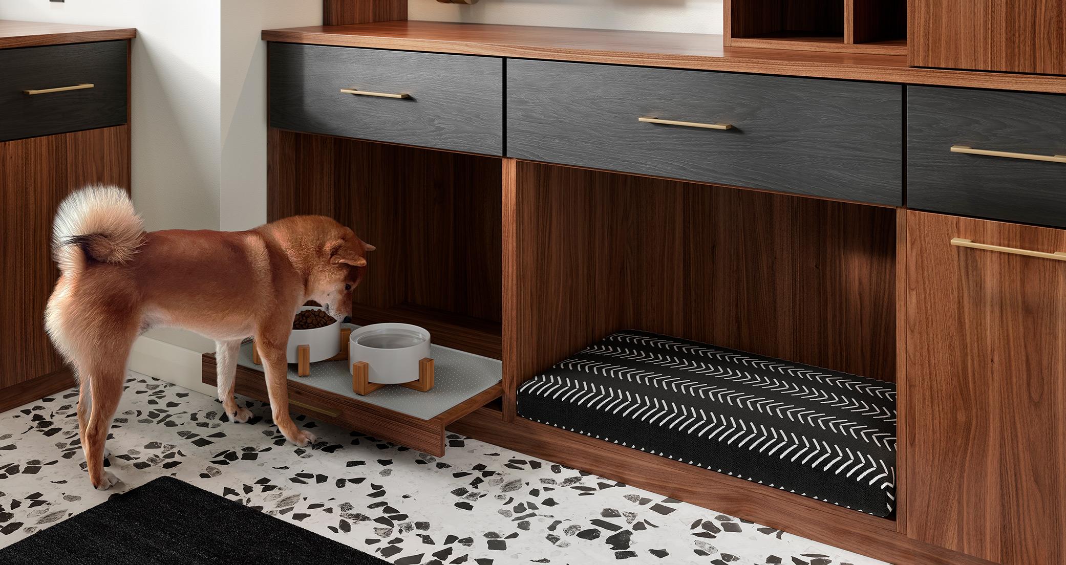 Una estación de alimentación integrada con cama para mascotas diseñada por California Closets, que incluye un gabinete de madera con cajones negros, una bandeja extraíble con tazones de comida y agua, y un área de descanso acogedora para mascotas. | California Closets