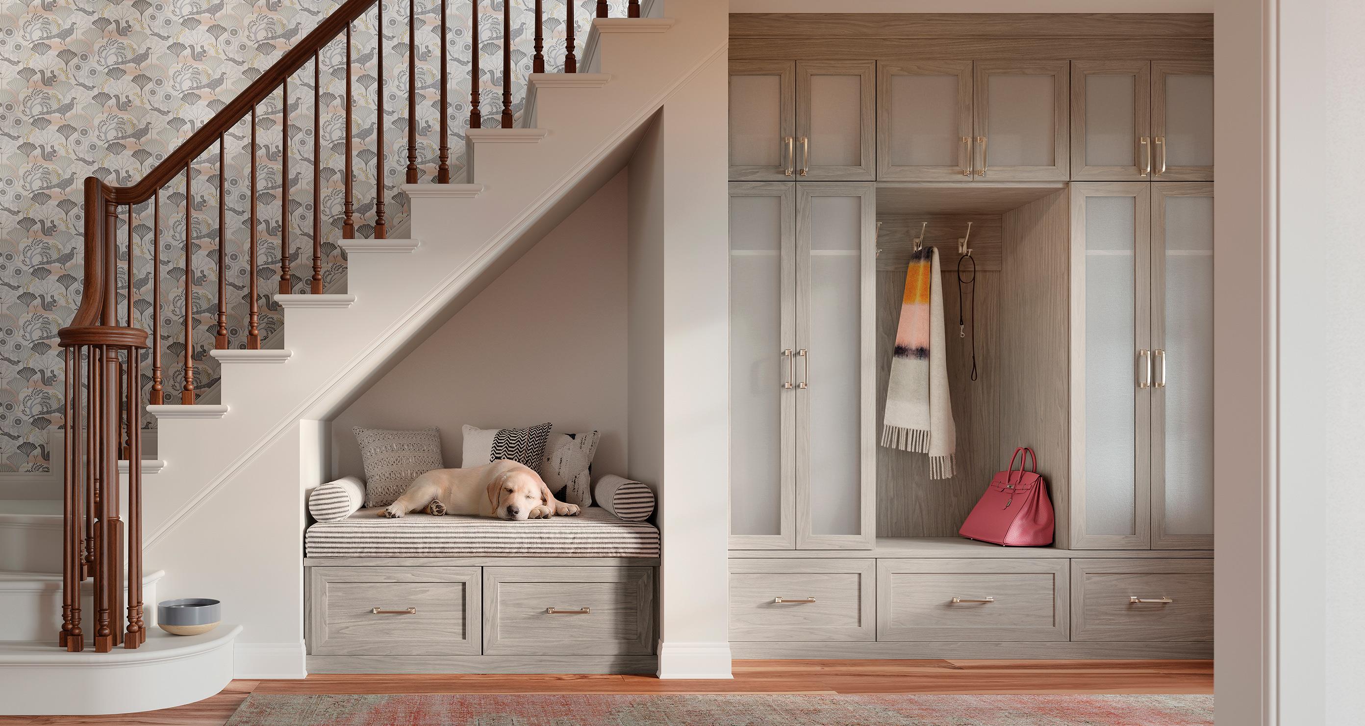 Un diseño acogedor de almacenamiento bajo escalera por California Closets que incluye un banco con cojines y un perro dormido, junto a gabinetes empotrados y espacio para colgar abrigos y accesorios.