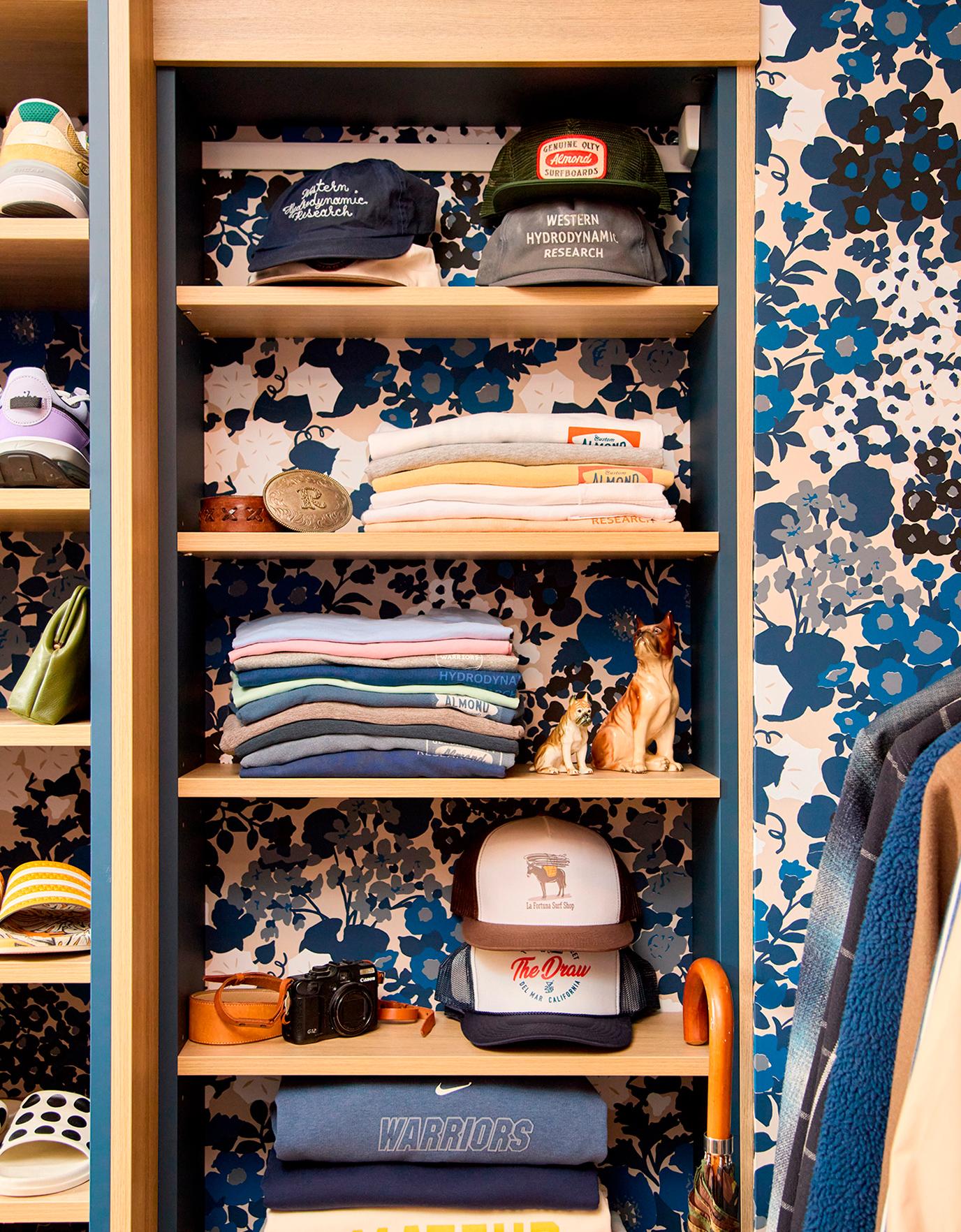 Un armario bellamente organizado por California Closets con estantes que muestran sombreros, ropa doblada, una cámara y artículos decorativos sobre un fondo de papel tapiz floral azul. | California Closets