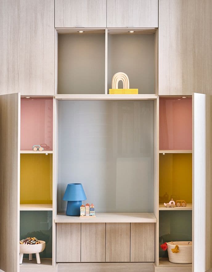 Light wood custom storage unit for a children’s room featuring integrated puck lighting and multi-colored back-painted glass panels in shades of blush, gold, and seafoam green.
