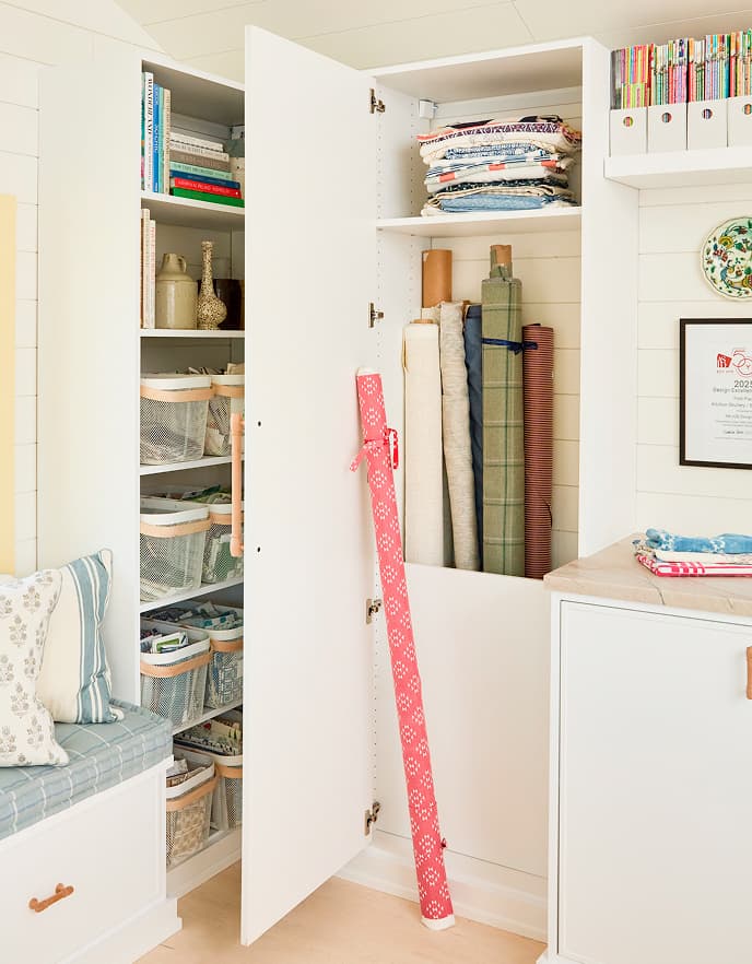 Close-up of custom California Closets storage in a design studio featuring tall cabinets for fabric bolts, open shelving with organized bins for swatches, and a white Dove finish with leather-pull hardware.