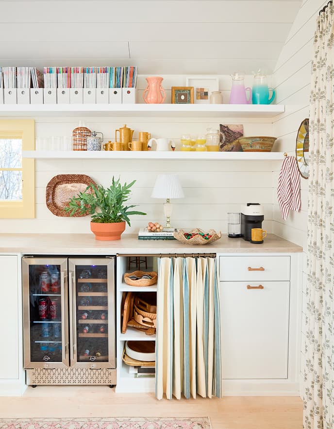 Una cocineta de estudio personalizada de California Closets que cuenta con un refrigerador para bebidas empotrado, gabinetes con acabado blanco Dove y tiradores de cuero, y estanterías abiertas flotantes que exhiben una colección de revistas y cerámicas decorativas.