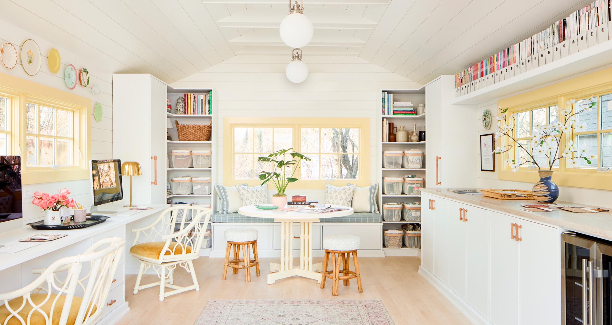 Una cochera independiente y luminosa convertida en un estudio de diseño profesional por California Closets, con gabinetes personalizados en blanco Dove, estaciones de trabajo dobles, un banco acolchado junto a la ventana y estanterías abiertas organizadas para muestras de diseño de interiores.