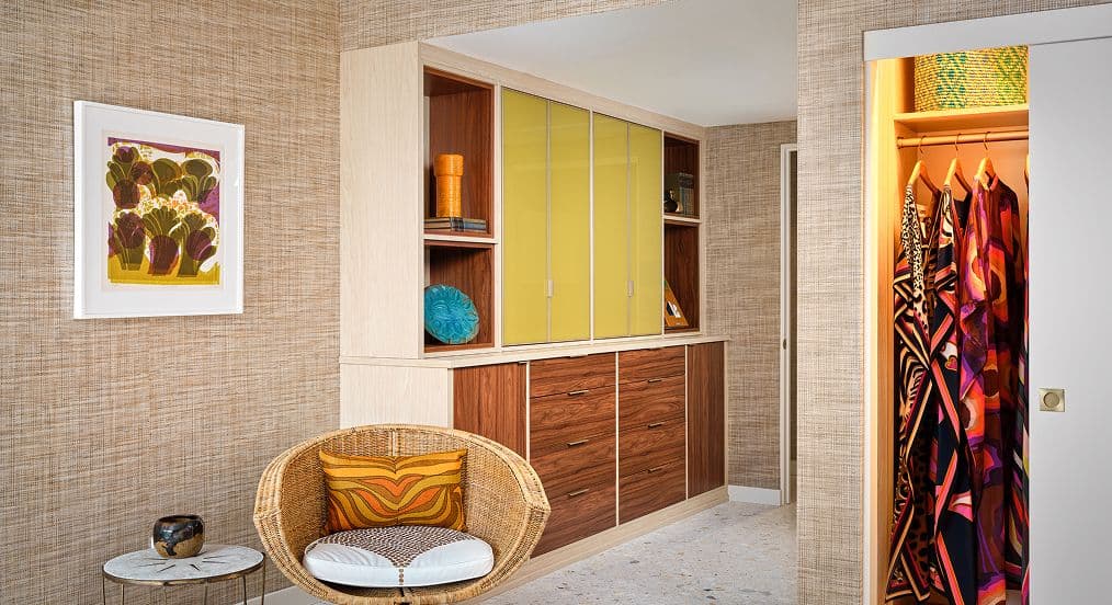 Custom bedroom storage suite featuring grasscloth wallpaper, a wicker accent chair, and a wood grain wardrobe unit with vibrant yellow back-painted glass door inserts.