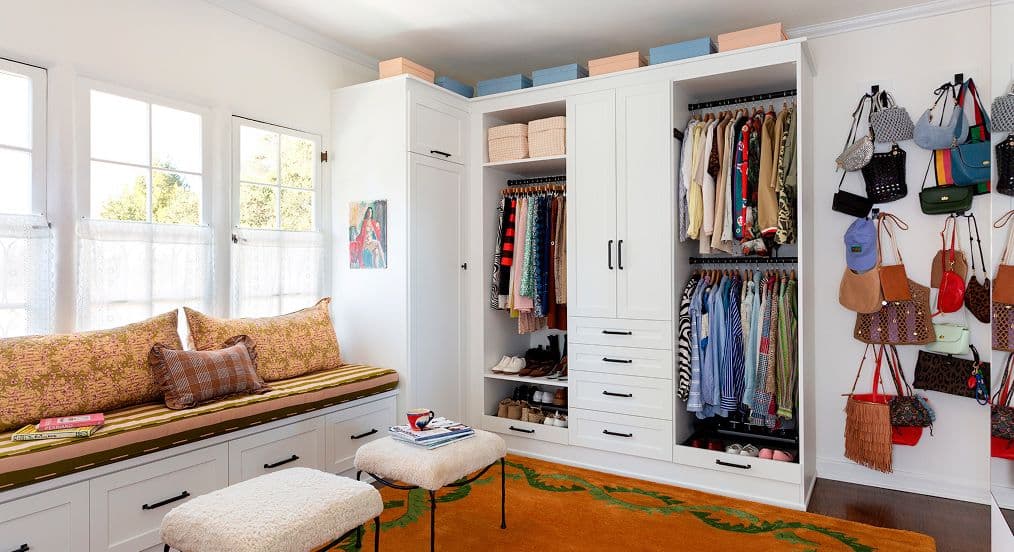 Vestidor a medida con ropa a la vista, ganchos para bolsos, gabinetes blancos y banco junto a la ventana de California Closets